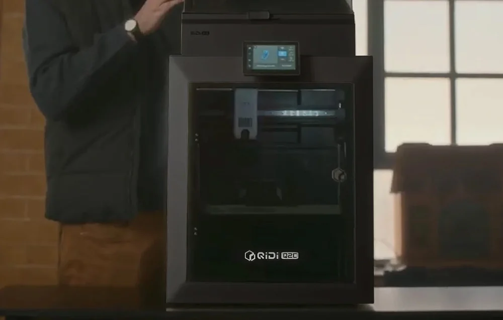 Nowoczesna czarna drukarka 3D QIDI Q2C na biurku, w tle pracujące osoby w biurze projektowym.